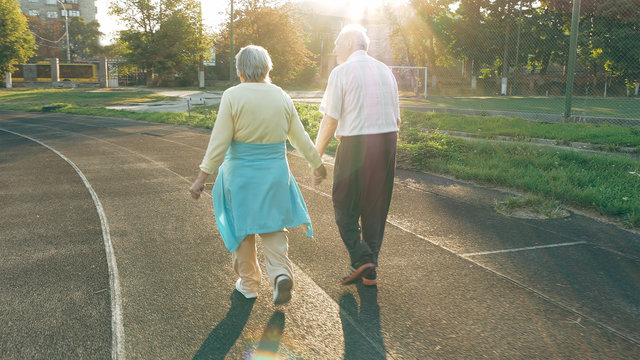 Senior couple taking a walk along the running track in summer. Healthy retirees enjoying morning walking on the stadium with camera lens flare.