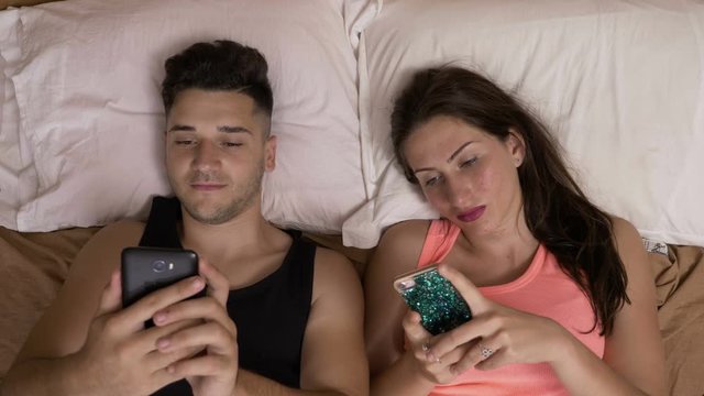 Addicted to smartphone couple surfing on internet watching on each other partner phone laying in bed in pajamas