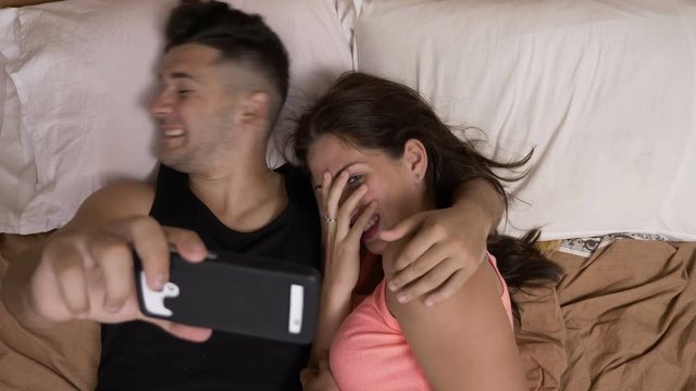 Girlfriend and boyfriend sitting comfortable in bed watching horror movie on internet using smartphone