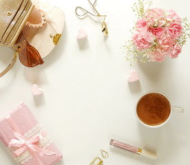 Feminine desk with bouquet of pink carnations in a vase, cup of cocoa , accessories, gift in pink packing and pink hearts on a white background. Holiday concept. Top view. copy space