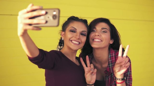 Two attractive women posing while taking selfie and showing peace sign. Beautiful girls taking photos with smart phone standing by the yellow wall. Slowmotion shot