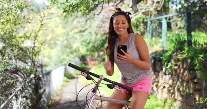 Trendy Hispanic woman sitting on bicycle outdoors, waving to someone off camera and smiling. Friendly Latina female resting on bike trail, messaging on cellphone on summer day. 4k 