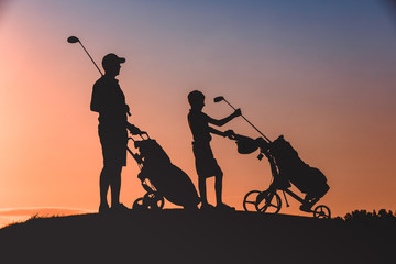 silhouettes of man with his son golfers walking with bags on golf course at sunset