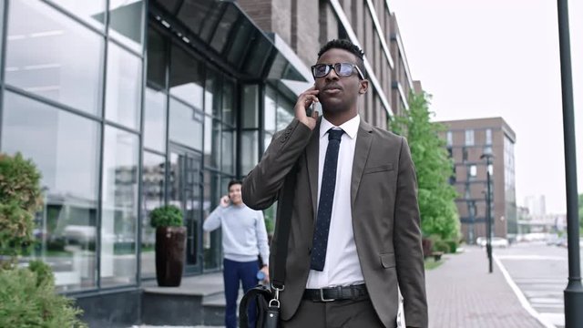 African businessman walking outdoors along modern office building in downtown and speaking on mobile phone