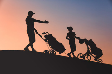 Fototapeta na wymiar silhouettes of man with his son golfers walking with bags on golf course at sunset