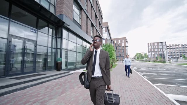 Slowmo of African businessman walking on street in downtown with suitcase and talking on mobile phone