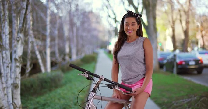 Portrait of smiling Latina female in tank top and shorts posing with bicycle outdoors. Trendy Hispanic woman sitting on bike near busy street, looking at camera with friendly expression. 4k 