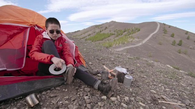 Young Asian woman hiker sitting in tent at campsite in mountains and fixing her broken boot with adhesive tape