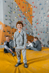 Smiling little boy in sportive attire standing in front of a climbing wall and looking at camera