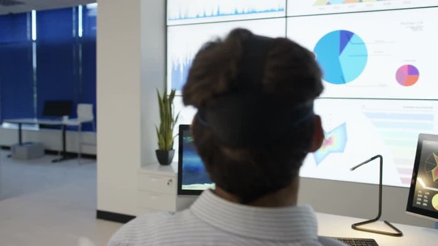  Businessman interacting with a virtual reality headset in futuristic office, computer screens showing financial information & world map