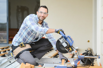 handsome young man carpenter using a circular saw while installing wood floor terrace outdoor in new house construction site