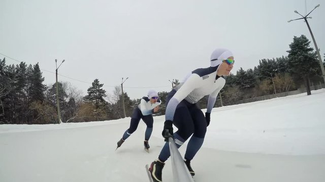 Shot from selfie stick held by female speed skater in professional sportswear and protective eyewear racing against teammate during practice in outdoor ice rink in winter