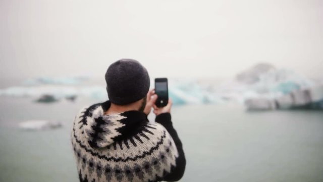 Young stylish man standing on the shore in Jokulsalon ice lagoon in Iceland and taking photos on smartphone.