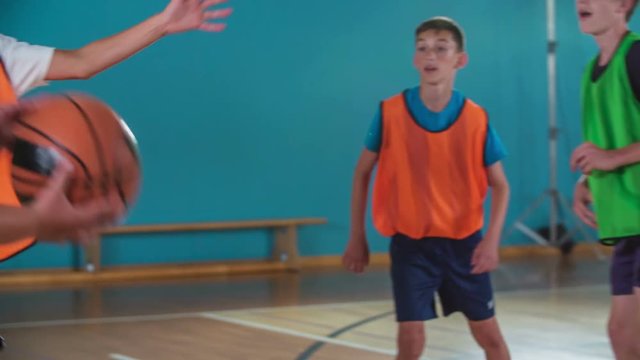 Orange and green teams are playing basketball with each other in a school gym. There are only boys. Secondary school.