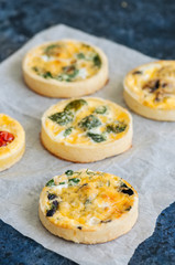 Set of savory mini tarts on a baking paper. Vegetable quiches with tomatoes, mushrooms, herbs, broccoly. Blue stone background. Close up and copy space.
