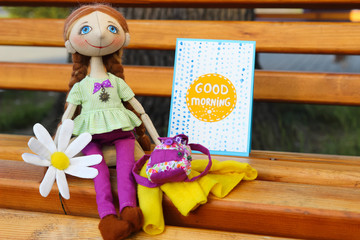 Textile handmade doll toy girl with blue eyes and a flower of a camomile sits on bench near postcard with inscription "Good morning"