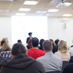 Business and entrepreneurship symposium. Female speaker giving a talk at business meeting. Audience in conference hall. Rear view of unrecognized participant in audience. Copy space on whitescreen.