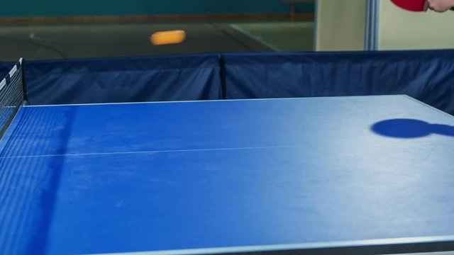 A small ball is going from one side of the table to another. Children are playing table tennis in a school gym.