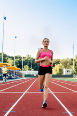 Sporty fitness woman jogging on red running track in stadium. Training summer outdoors on running track line with green trees on background.