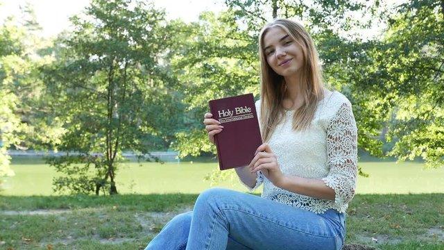4k.Young girl show Bible in  summer park  and smile.Christian  belief team. 