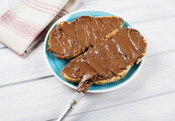 Chocolate spread on bread on a blue plate next to a knife and a napkin on wooden table.