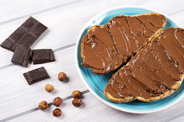 Chocolate spread on bread on a blue plate next to pieces of chocolate and hazelnuts on wooden table.
