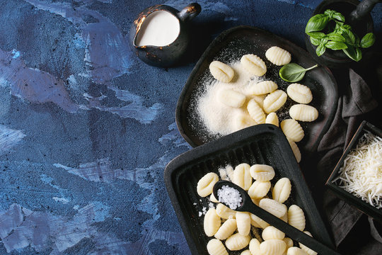 Raw uncooked potato gnocchi in black wooden plates with ingredients. Flour, grated parmesan cheese, basil, salt, jug of cream over dark blue concrete background. Top view, copy space Home cooking.