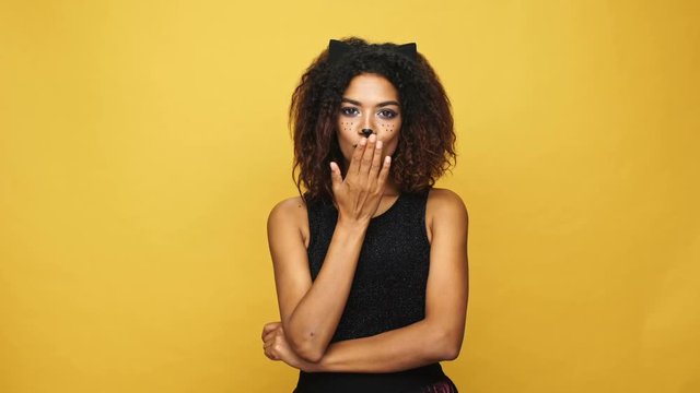 Beautiful afro american lady with cat make-up and ears flirting and calling you isolated over yellow wall