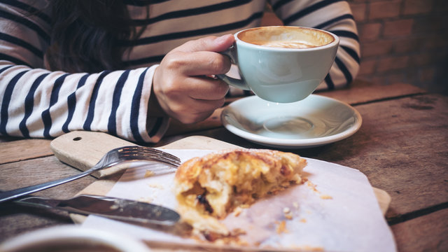 Closeup image of a woman holding and drinking hot latte coffee with a piece of raisin danish on wooden vintage table in coffee shop