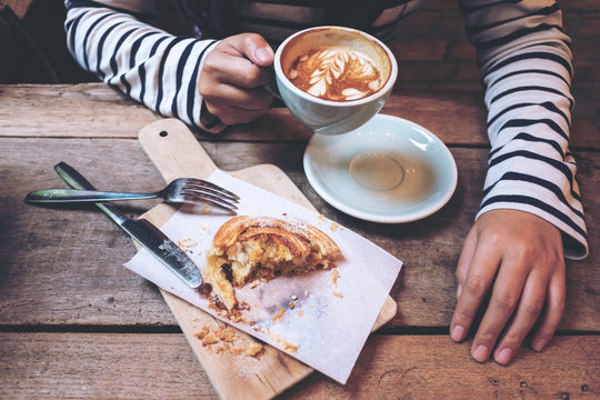 Top view image of a woman holding and drinking hot latte coffee with a piece of raisin danish on wooden vintage table in coffee shop