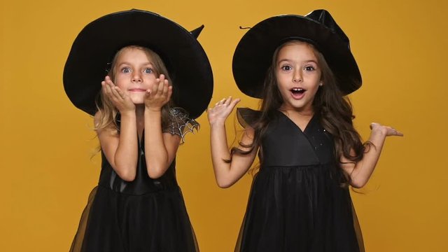 Little playful girl in witch dresses and hats hide their faces and surprising isolated over orange