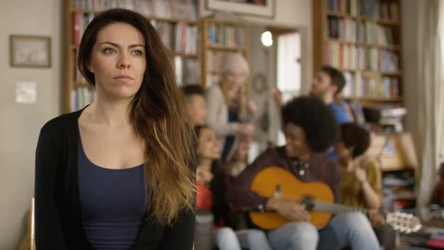 Young woman in shared apartment feeling sad and isolated while others socialize in background.