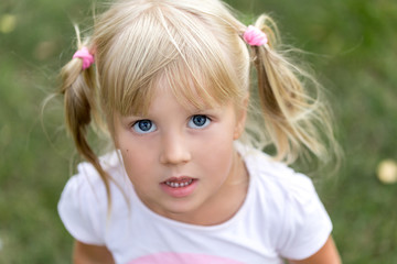 Close up portrait of cute adorable little blonde caucasian girl in a white T-shirt in a park, looking aside, thinking or dreaming of something, puzzled or astonished, happy childhood concept. - Powered by Adobe