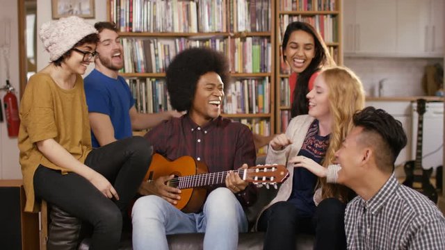 Happy young friends in shared apartment playing guitar and socializing.