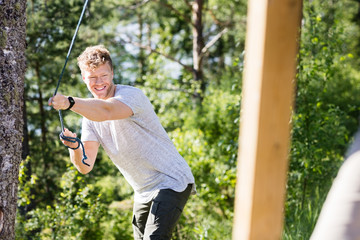Happy Man Pulling Rope Attached To Wooden Structure In Forest