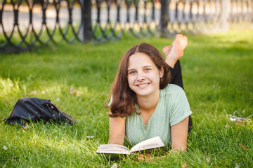 A cute teen girl lies on a green lawn, smiling and looking at the camera outdoors. A student is reading a book in the park, lying on the grass in the park