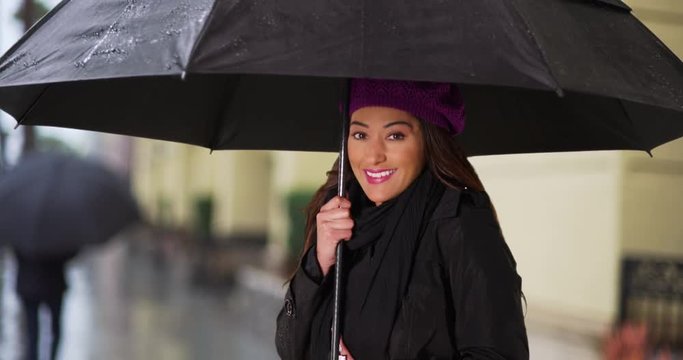 Cute Latina female dressed in raincoat holding black umbrella outdoors. Portrait of young woman shivering outside in the cold weather, smiling at camera. 4k 