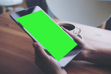 Mockup image of business woman's hands holding black tablet pc with blank green screen and coffee cup on wooden table in cafe background