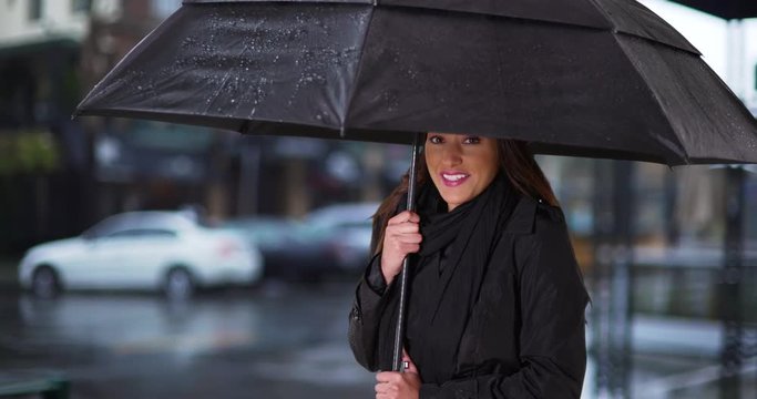 Cute Latina female taking shelter from the rain with black umbrella. Portrait of young woman standing under umbrella on gloomy day, smiling at camera. 4k 