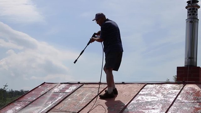 Man is washing the roof with a high pressure washer. Contrast silhouette of a man washing a metal sheet roof by water. 