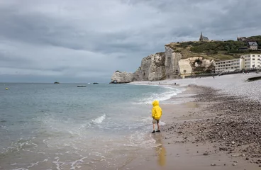 Fototapeten Child in yellow raincoat on the beach of Etretat, France. The boy is standing on the beach and looking into the distance,. © Erik_AJV