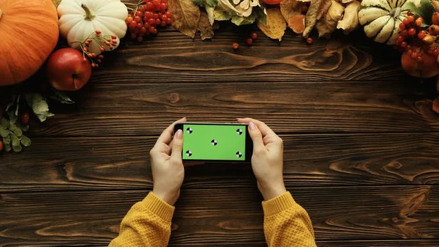 Top view of fall composition with pumkins, maple leaves, nuts and black smart phone on the brown wooden table. Woman holding phone with green screen. Chroma key. Tracking motion