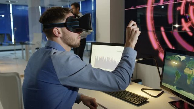 Businessman interacting with a virtual reality headset in futuristic office, computer screens showing financial information and animated graphics