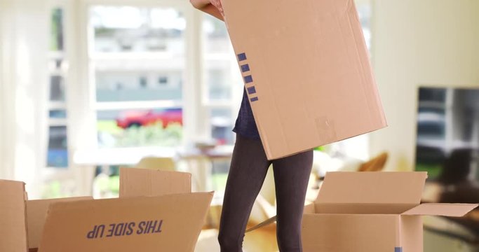 Young female carrying heavy cardboard box past camera inside living room. Casually dressed woman struggling with big moving box indoors. 4k 