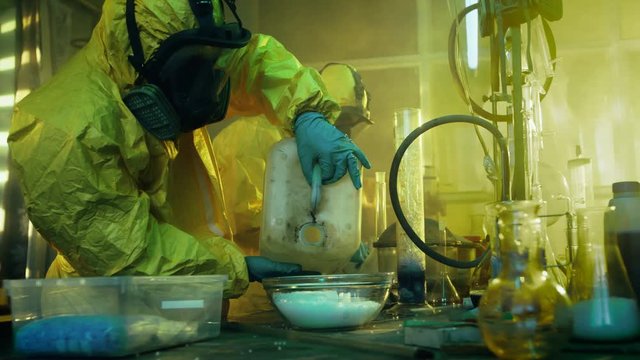 In the Underground Drug Laboratory Two Clandestine Chemists Covered in Protective Coveralls and Gas Masks Mix Chemicals to Synthesise Drugs. They Work in the Abandoned Building. 