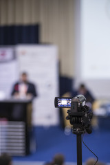 Back View of Compact Videocamera. Positioned Against Blurred Background with Host Speaking on Stage.