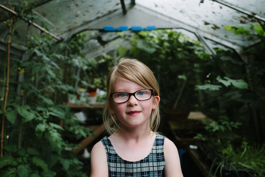 Portrait of young girl with long blonde hair and glasses standing in a greenhouse.