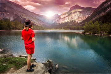Une jeune femme blonde de dos avec une robe rouge et un chapeau au bord d'un lac de montagne sous un coucher de soleil
