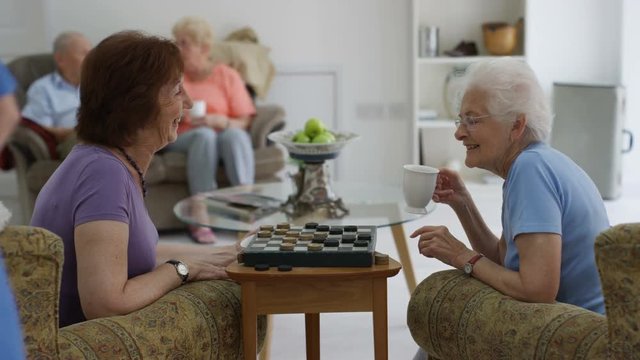 Cheerful senior ladies playing draughts in a nursing home and young nurse taking care of residents.