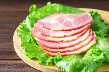 Sliced ham with fresh green lettuce leaves on a round cutting board. Meat products on a brown wooden table.